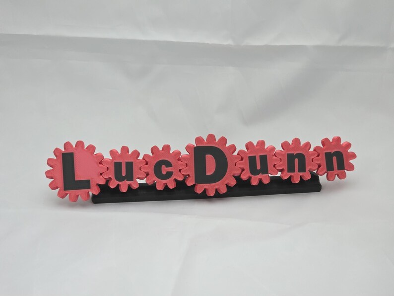 K&ouml;nnte beinhalten: Ein personalisiertes Namensschild mit dem Schriftzug LucDunn in schwarzen Buchstaben, wobei jeder Buchstabe auf einer rosafarbenen, zahnradf&ouml;rmigen Basis zentriert ist. Die Zahnr&auml;der sind horizontal verbunden und auf einem schwarzen Sockel montiert.