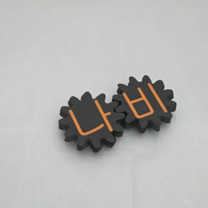 K&ouml;nnte beinhalten: Zwei ineinandergreifende schwarze zahnradf&ouml;rmige Objekte mit orangefarbenen koreanischen Schriftzeichen. Das linke Zahnrad zeigt das Zeichen '나' und das rechte Zahnrad das Zeichen '비', was ein stimmiges mechanisches Design mit matter Oberfl&auml;che ergibt.