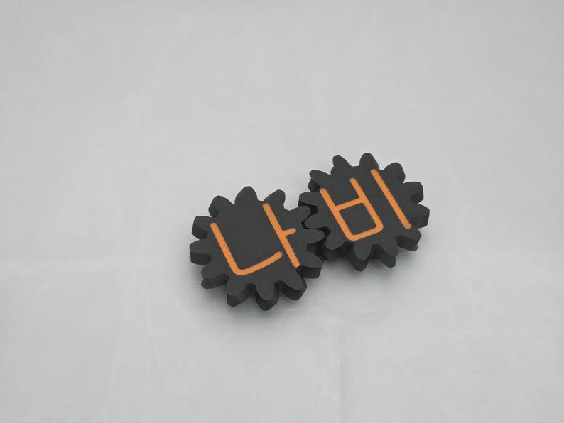 K&ouml;nnte beinhalten: Zwei ineinandergreifende schwarze zahnradf&ouml;rmige Objekte mit orangefarbenen koreanischen Schriftzeichen. Das linke Zahnrad zeigt das Zeichen '나' und das rechte Zahnrad das Zeichen '비', was ein stimmiges mechanisches Design mit matter Oberfl&auml;che ergibt.