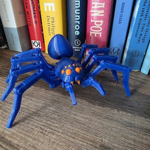 May include: A blue plastic spider toy with orange spots on its body and eight articulated legs. The spider is sitting on a wooden surface.