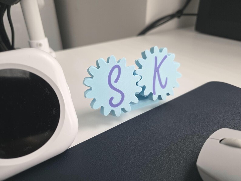K&ouml;nnte beinhalten: Ein hellblaues Schreibtischornament bestehend aus zwei ineinandergreifenden zahnradf&ouml;rmigen Teilen. Jedes Zahnrad hat einen violetten Buchstaben in der Mitte aufgedruckt, wobei der Buchstabe S auf dem linken und der Buchstabe K auf dem rechten Zahnrad steht.
