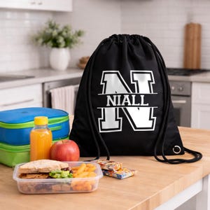 May include: Black drawstring bag featuring the name "NIALL" in white block letters. The bag is on a wooden surface with a packed lunch, including a sandwich, apple, juice, and snacks. A blue and green lunch bag is also present.