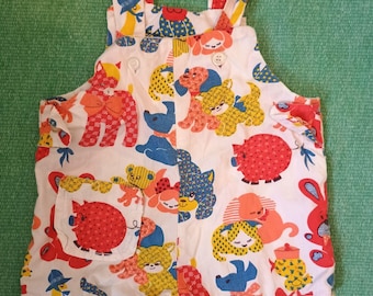 May include: White baby overalls with a colorful pattern of cartoon animals, including dogs, cats, and pigs. The overalls have adjustable straps and a small pocket on the front. The background is a solid green.