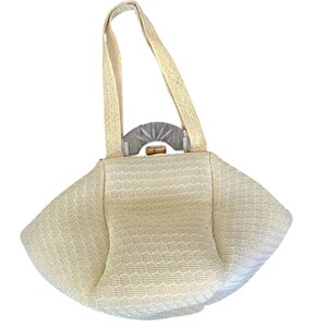 May include: A cream-colored woven handbag with a structured, fan-like silhouette. The bag features two matching woven top handles and a semi-circular, translucent gray plastic accent piece positioned above a small, horizontal wooden clasp closure.