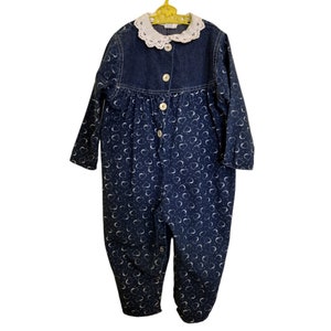 May include: A blue denim jumpsuit with a white lace collar and a heart print. The jumpsuit has long sleeves and buttons down the front.