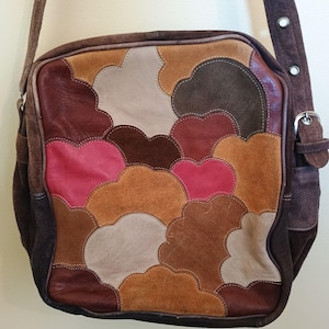 以下が含まれることがあります： 茶色の革製ショルダーバッグ、パッチワークデザイン。茶色、タン、ピンクの色合いの、アースカラーのスエードと革のさまざまな部分が、雲のようなパターンで縫い合わされています。バッグには、長さ調節可能な長いストラップが付いています。