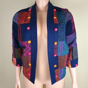 May include: A navy blue open-front jacket featuring a patchwork design with various geometric patterns in shades of orange, pink, and teal. The garment has three-quarter length sleeves and is accented with small, round, colorful decorative buttons.