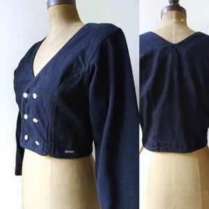May include: A black cropped denim jacket with a v-neck, long sleeves, and a button closure. The jacket has a Bongo logo on the bottom right side.