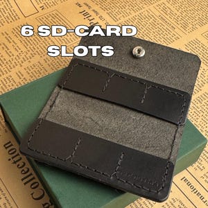 Pode incluir: Suporte de cartão SD em couro preto com seis ranhuras, aberto para mostrar o interior. Está em cima de uma caixa verde, com um fundo de jornal. O texto "6 SD-CARD SLOTS" é visível.