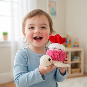 May include: A crocheted turtle toy with a cupcake design. The cupcake has a pink base, white frosting, and two red heart decorations. The turtle is cream-colored with a smiling face and is held by a smiling child.