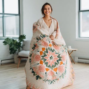 May include: A cream-colored quilt with a floral design featuring pink and orange flowers and green leaves. The quilt has a central starburst pattern and is surrounded by a border of floral elements. The quilt is displayed in a well-lit room.
