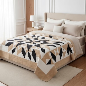 May include: A bed with a geometric patterned quilt. The quilt features black, white, and tan diamond and triangle shapes. The bed is made up with neutral-colored pillows and a beige bed skirt. A matching throw is folded over the edge of the bed.