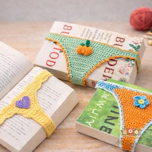 Könnte beinhalten: Drei gehäkelte Buchumschläge in Form von Unterwäsche, jeweils mit einzigartigen Designs. Einer ist gelb mit einem lila Herz, ein anderer ist grün mit einem orangefarbenen Kürbis, und der dritte ist orange mit einer blauen Blume. Die Umschläge liegen auf Büchern.