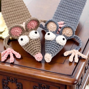 Peut inclure: Deux écharpes en forme de souris crochetées, une marron et une grise. Chaque souris a de grands yeux blancs, des oreilles roses et un nez rose. Les écharpes sont posées sur une surface en bois.