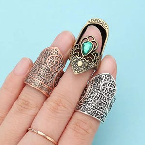 May include: Three metal rings with intricate filigree patterns. One ring is copper-toned, one is silver-toned, and the third is gold-toned with an ornate open-top design featuring a central teardrop-shaped green gemstone accent.