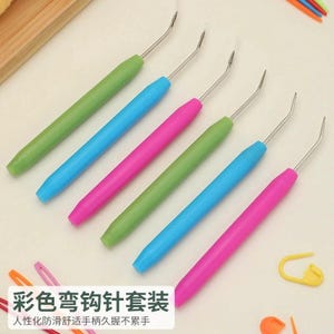 Peut inclure: Un ensemble de six outils de loisirs créatifs avec des manches colorés verts, bleus et roses, chacun doté d'un crochet métallique fin et incliné à l'extrémité. Le texte sur l'image indique "彩色弯钩针套装" et "人性化防滑舒适手柄久握不累手".