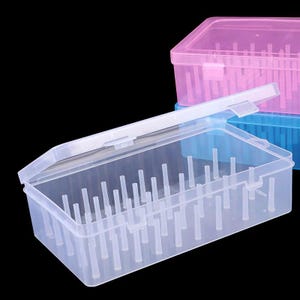 May include: Clear plastic storage box with an open lid, revealing internal vertical dividers. The box is rectangular and has a hinged lid. Other boxes in pink and blue are visible in the background. The boxes are designed for organizing small items.