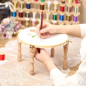Puede incluir: Un pequeño soporte de bordado redondo de madera con cuatro patas, que sostiene un bastidor de tela blanca. Una persona usa una aguja para bordar un diseño floral. Bobinas de hilo de colores en el fondo, sugieren un tema artesanal.