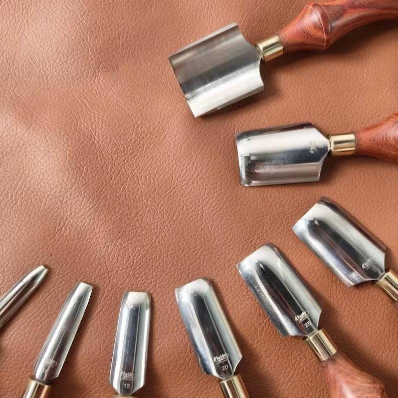 May include: A collection of leatherworking tools, including various gouges and a burnisher, arranged on a brown leather surface. The tools have polished metal blades and wooden handles with brass ferrules. The gouges have curved blades.