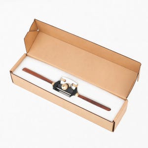 May include: A woodworking spoke shave tool with dark brown wooden handles, a black metal body, and a polished silver blade, resting inside a rectangular cardboard box lined with white foam padding for secure storage and protection.
