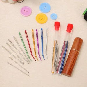 May include: A collection of sewing needles in various colors and sizes, along with colorful buttons. The needles are in clear tubes with red caps and a wooden needle case. Buttons are pink, blue, and yellow.