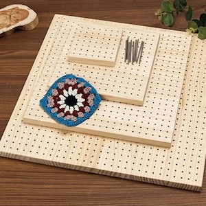 May include: Wooden crochet blocking boards with evenly spaced holes, a blue, red, and white crochet square, and metal pins. The boards are arranged in a layered fashion, showcasing a craft project in progress.