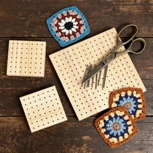 May include: Three light-colored wooden blocking boards with grid holes, a pair of metal scissors, several thin metal pins, and three crocheted granny squares in blue, white, brown, and yellow tones arranged on a dark wooden surface.