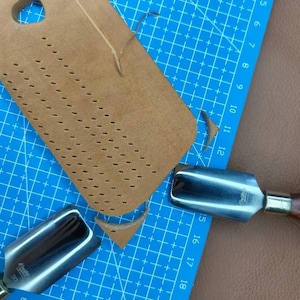 May include: A close-up shot of leather crafting tools and materials. A piece of tan leather with evenly spaced holes sits on a blue cutting mat. Two metal tools with wooden handles are also visible, suggesting leatherwork or crafting.