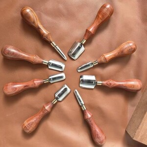 May include: A collection of leatherworking tools arranged in a circular pattern. The tools feature polished metal blades and wooden handles with a rich, reddish-brown hue. The handles are intricately carved, and the tools are set against a brown leather surface.