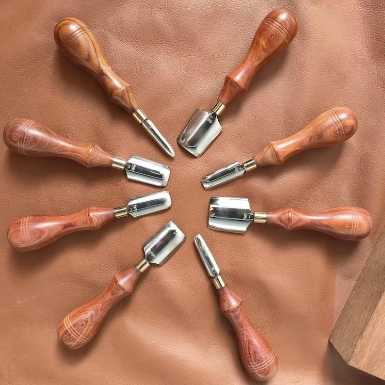 May include: A collection of leatherworking tools arranged in a circular pattern. The tools feature polished metal blades and wooden handles with a rich, reddish-brown hue. The handles are intricately carved, and the tools are set against a brown leather surface.