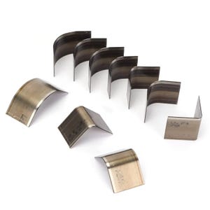 May include: A collection of metal corner cutters, arranged on a white surface. The cutters are various sizes and shapes, with a curved or right-angle design. The metal has a silver-gray color, and some cutters have markings.