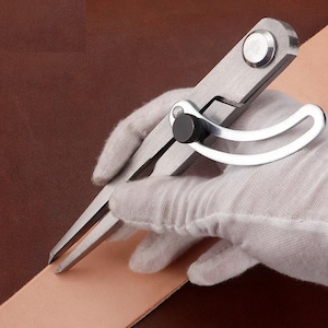 May include: A stainless steel wing divider tool with two pointed legs and a curved adjustment arm featuring a black locking knob. The metal tool is positioned against a strip of light tan leather to mark a straight line along the edge.
