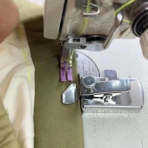 May include: A metal sewing machine presser foot attachment with a silver finish is positioned on a white machine bed. The device guides a strip of light purple fabric tape onto a piece of olive green fabric as the needle stitches the material.