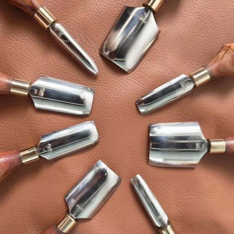 May include: A collection of leatherworking tools arranged in a circular pattern. The tools have polished metal blades, wooden handles, and brass ferrules. The background is a brown leather surface.