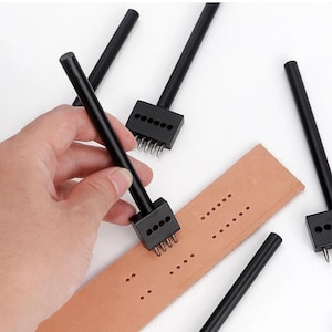 May include: A hand using a black leather punch tool to create holes in a tan leather strip. Several other black leather punches are scattered around the leather strip. The tools have a black handle and a rectangular head with multiple prongs.