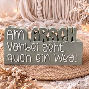 Könnte beinhalten: Ein dekorativer Block in Salbeigrün mit dem deutschen Satz "Am Arsch vorbei geht auch ein Weg!" in weißen Buchstaben. Der Block steht auf einer gewebten Matte mit getrockneten floralen Akzenten, mit einem Makramee-Wandbehang im Hintergrund.
