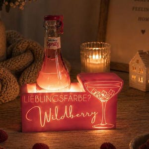 Könnte beinhalten: Eine beleuchtete rote Box mit dem Text "LIEBLINGSFARBE? Wildberry" und einem Cocktailglas-Design. Eine Flasche Spritz steht obenauf. Himbeeren und Johannisbeeren sind verstreut. Im Hintergrund befinden sich eine Kerze und ein gerahmtes Schild.