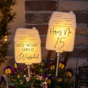 Puede incluir: Dos estacas de jardín iluminadas con forma de casa en una cesta de mimbre llena de flores moradas y amarillas. Una estaca dice "HIER WOHNT FAMILIE Westphal" y la otra "Haus Nr. 15". Un farol de metal oscuro se encuentra junto a la cesta.