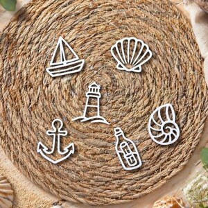 Könnte beinhalten: Ein runder, gewebter Tischset mit weißen maritimen Ausschnitten. Die Ausschnitte umfassen ein Segelboot, eine Muschel, einen Leuchtturm, einen Anker, eine Flasche und eine Nautilus-Schale. Das Tischset ist von Sand und Muscheln umgeben.