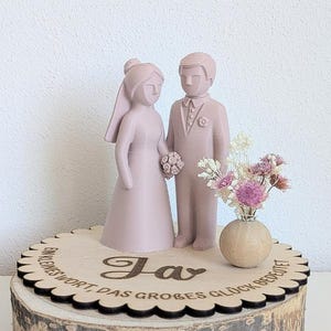 Könnte beinhalten: Ein hellrosa Tortenaufsatz für Hochzeiten mit einer Braut- und Bräutigamfigur auf einem runden Holzsockel. Der Sockel ist mit dem Text "Ja, ein kleines Wort, das großes Glück bedeutet" graviert und enthält eine kleine Holzvase mit getrockneten Blumen.