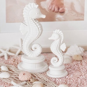 May include: Two white seahorse figurines of different sizes, each featuring a textured, segmented body and a curled tail. Both figures are mounted on circular, tiered bases and are crafted with a smooth, matte finish, displaying detailed fins.