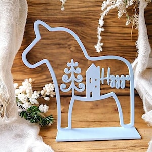 May include: A light blue wooden Dala horse figurine with a cutout design featuring a tree, house, and the word "Happy". The figurine stands on a rectangular base and is set against a wooden background.