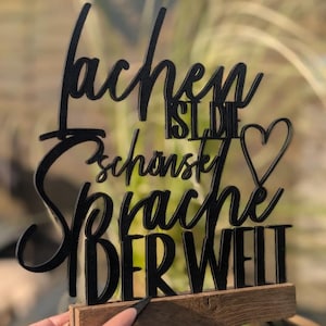 May include: Black wooden sign with the German phrase "Lachen ist die schönste Sprache der Welt" (Laughing is the most beautiful language in the world). The sign is mounted on a small wooden base and features a heart symbol.