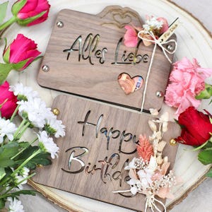 May include: Two wooden boxes with cut-out designs. The top box has the text "Alles Liebe" and a heart-shaped cutout. The bottom box has the text "Happy Birthday". Both boxes are decorated with dried flowers.