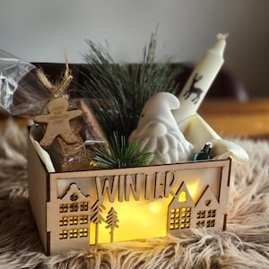 May include: A wooden box with the word "WINTER" cut out, illuminated by small lights. The box contains a white gnome figurine, a candle with a deer silhouette, a gingerbread man ornament, and greenery. The box sits on a fluffy, brown surface.