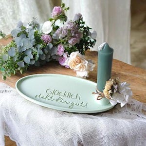 May include: A light green oval tray with the text "Gl&uuml;cklich steht dir gut!"  A teal candle sits on the tray with dried flowers and twine around the base.