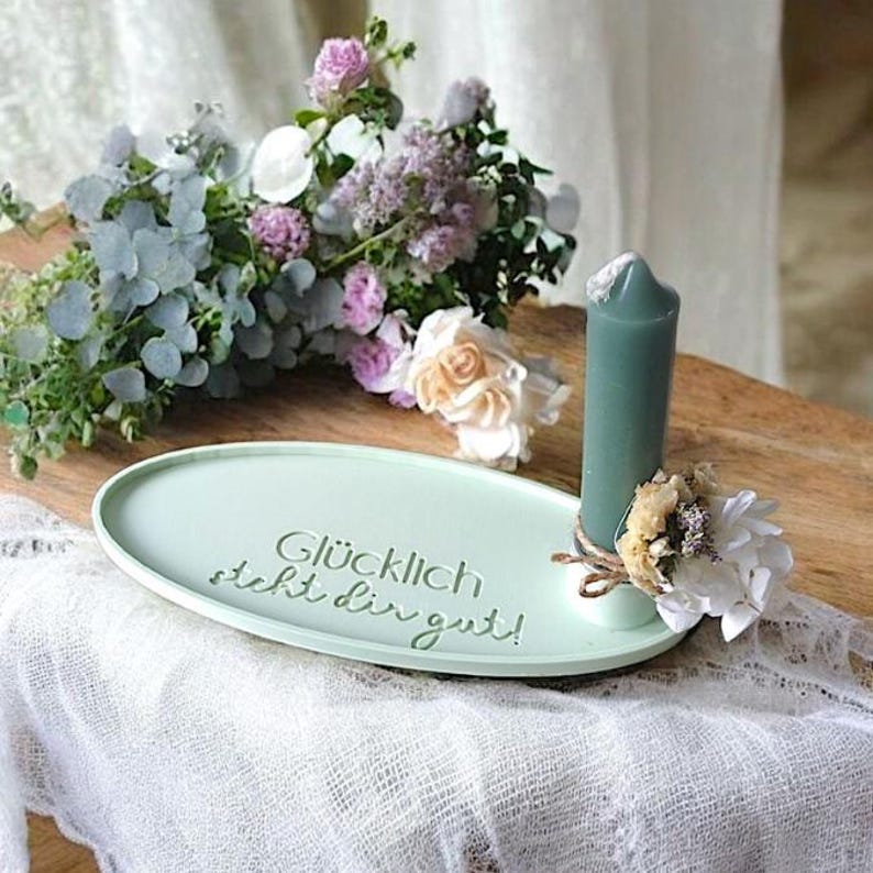 May include: A light green oval tray with the text "Gl&uuml;cklich steht dir gut!"  A teal candle sits on the tray with dried flowers and twine around the base.