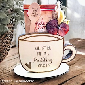 Puede incluir: Una caja de regalo de madera con forma de taza con un platillo, con el texto "WILLST DU MIT MIR Pudding Löffeln?" y un corazón. Incluye una cuchara de madera y un paquete de mezcla para pudin, con hojas decorativas y flores secas.