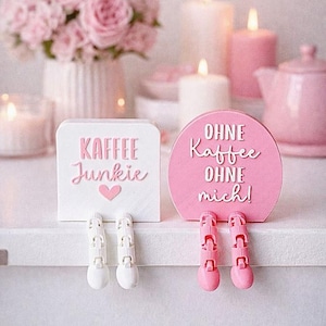 May include: Two decorative signs with legs. One is white with pink text that reads "KAFFEE Junkie" and a heart. The other is pink with white text that reads "OHNE Kaffee OHNE mich!". The background includes candles and flowers.