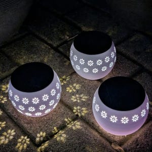 May include: Three spherical solar lights with a white finish and a repeating cutout flower pattern. Each light features a flat black circular solar panel on top and emits a soft glow that projects floral shadows onto the ground surface.